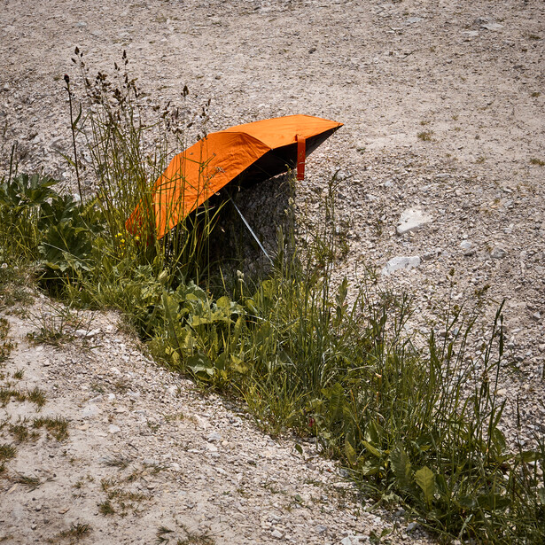 Orange umbrella lying on gravel ground beside sprouting weeds and dried plant stems