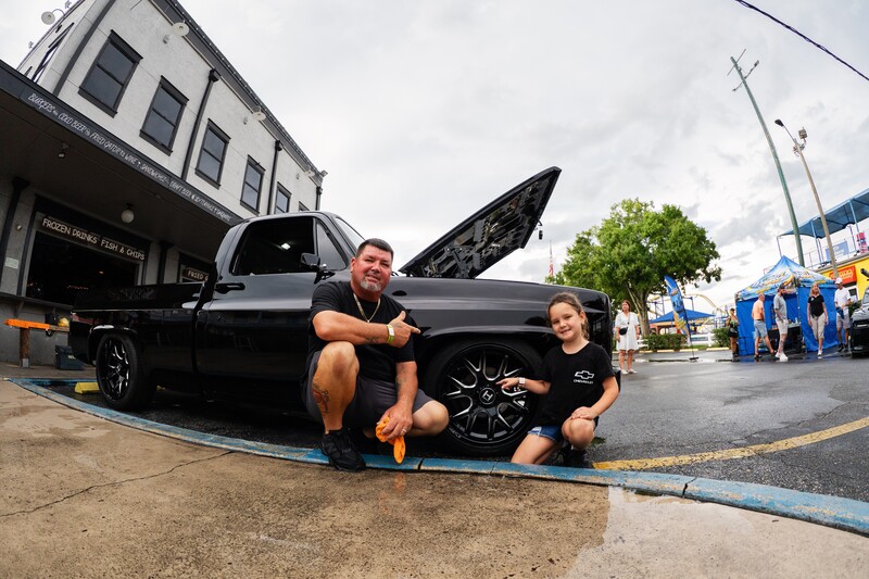 Two people crouching beside a lifted black pickup truck with hood open at a car meet