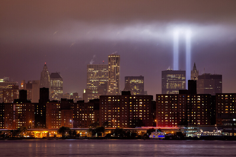Manhattan skyline at night with twin memorial light beams illuminating purple-toned sky