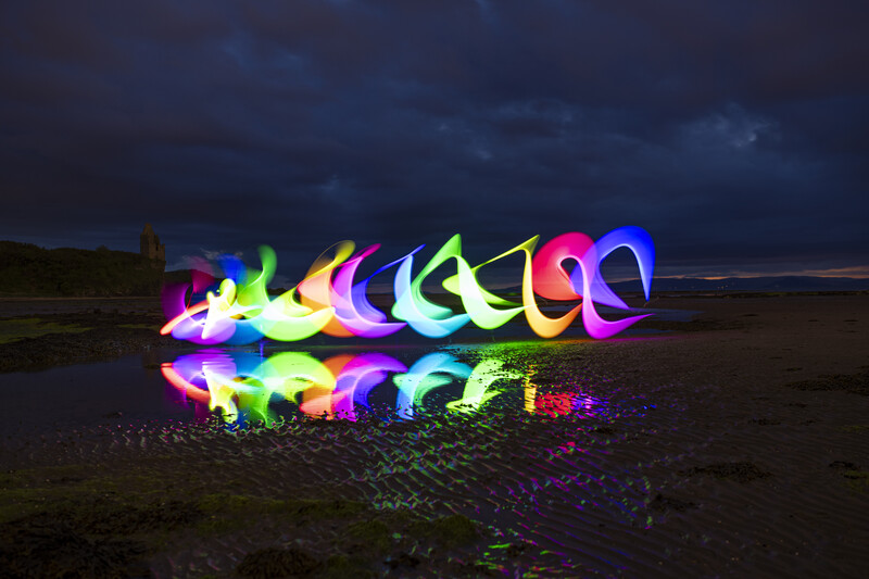 Neon light painting of abstract curved shapes glowing in rainbow colors on dark beach sand at dusk