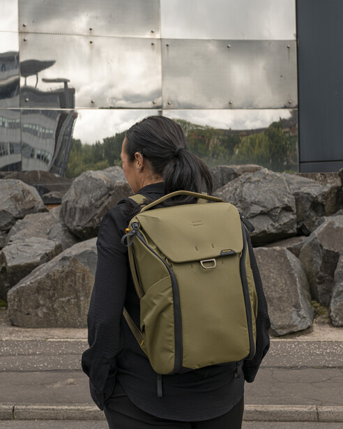 Person wearing olive-green backpack viewed from behind, standing in front of large rocks and billboard