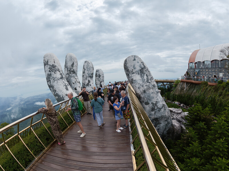Tourists walk across wooden bridge toward giant stone hands emerging from mountainside