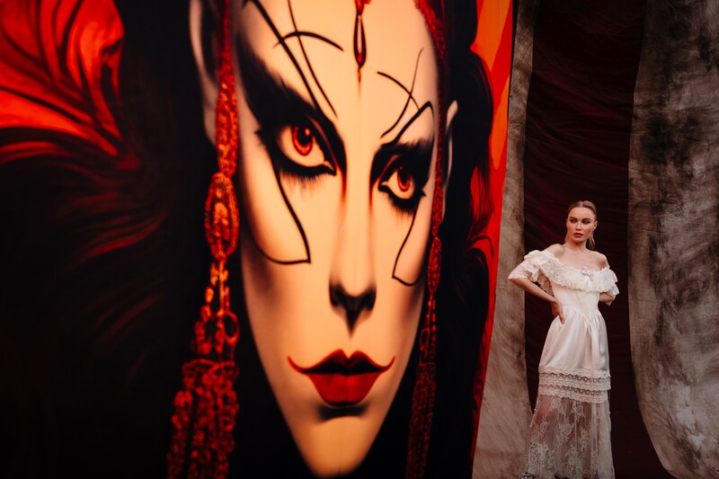 Woman in white off-shoulder dress standing beside large portrait projection with dramatic red and black makeup