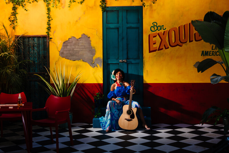 Woman in blue dress holding acoustic guitar seated against vibrant yellow and red wall
