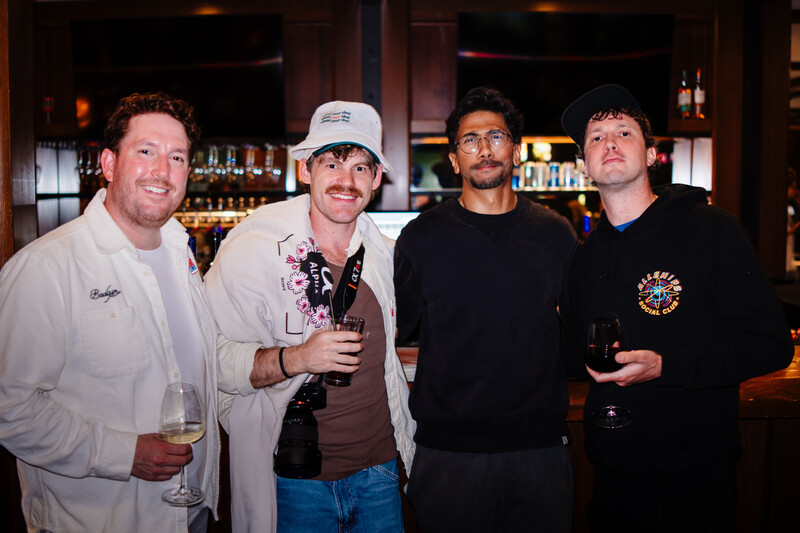 Four men posing together at an indoor bar, holding drinks and smiling at the camera