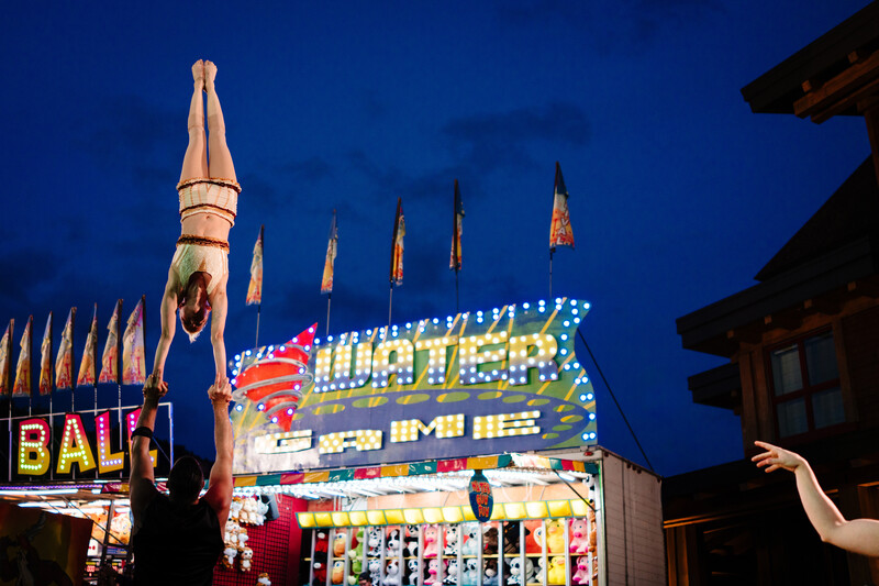Illuminated vintage carnival game booths and neon signs at dusk with lit spires in background