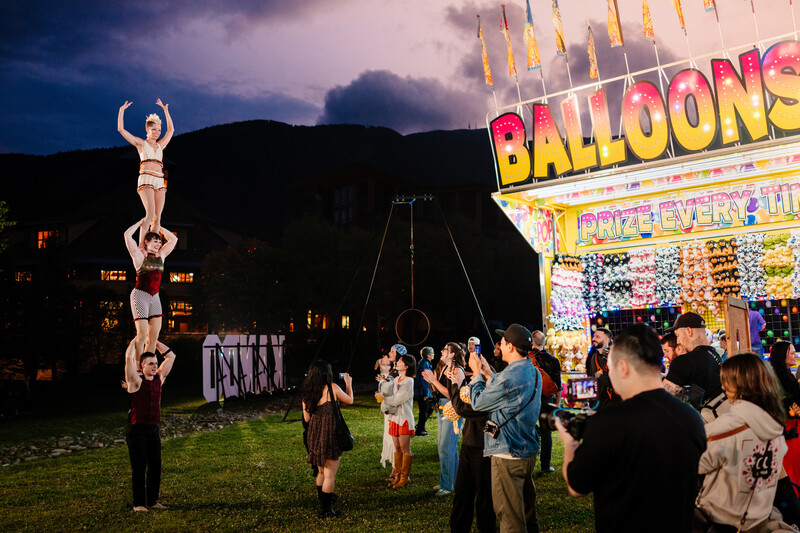 Acrobat balancing performers stacked vertically at dusk carnival with illuminated balloon game booth