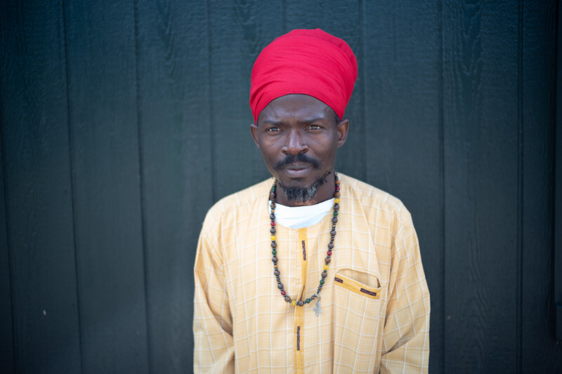 Man wearing a bright red head wrap and beaded necklaces against a dark wooden backdrop