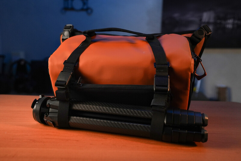 Orange waterproof dry bag with black straps displayed next to a rolled carbon fiber tripod