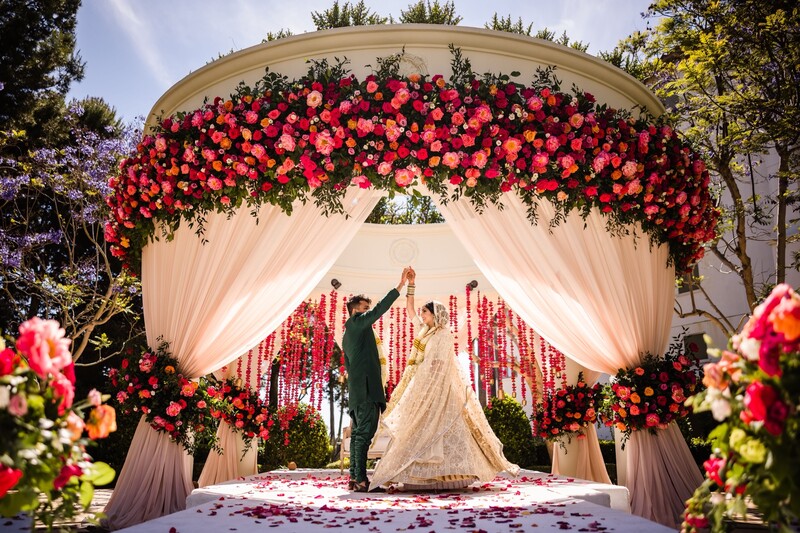 Bride and groom at wedding ceremony altar framed by red floral arch and cream drapes
