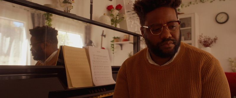 Man in mustard sweater and glasses sitting at piano with sheet music