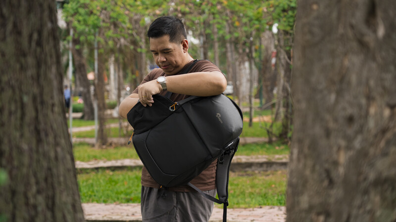 Man in black shirt holding camera bag in tree-lined park