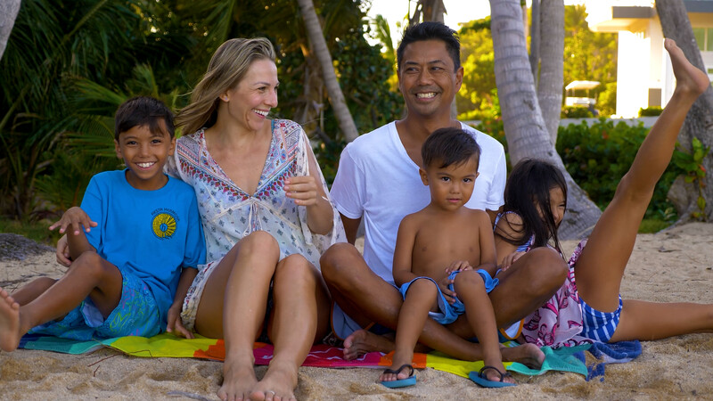 Family of five sitting together on sand at a beach with palm trees in background