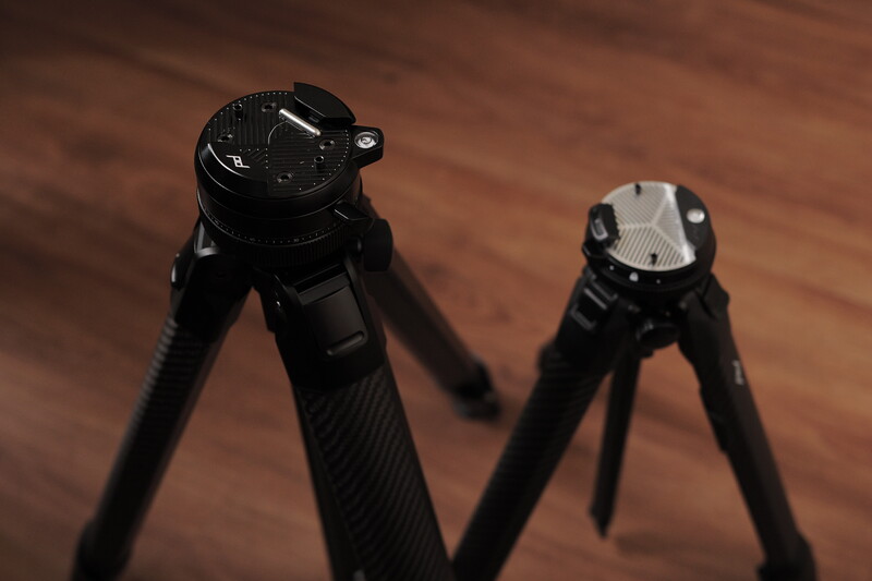 Two black rifle bipods with legs extended, displayed on a wooden surface