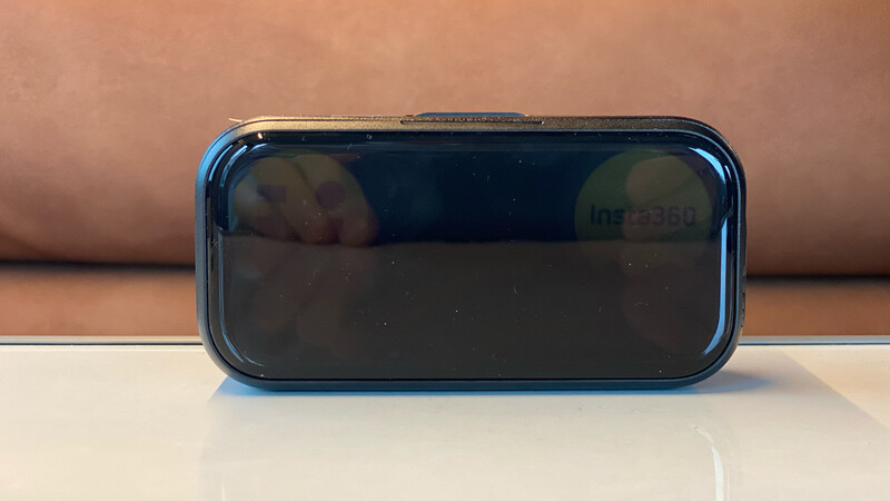 Compact black Insta360 device with a glossy rounded front panel, standing upright on a white reflective surface against a tan background.