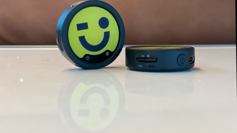 Two round black electronic devices on a glossy white surface. The device on the left shows a lime-green face with a dark blue winking smiley graphic.