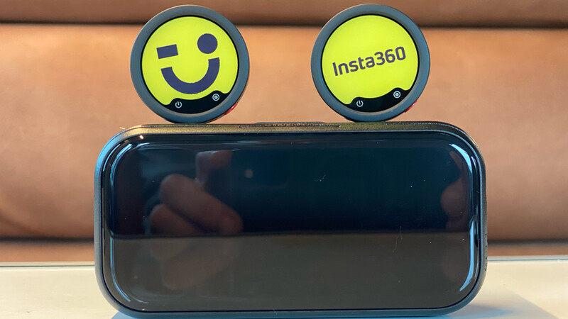 Two small round Insta360 cameras with yellow faces sit on top of a glossy black charging case against a tan leather couch.