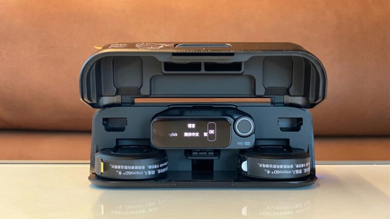 Open wireless microphone charging case showing a central receiver with a small screen and two clip-on transmitter mics docked on either side.