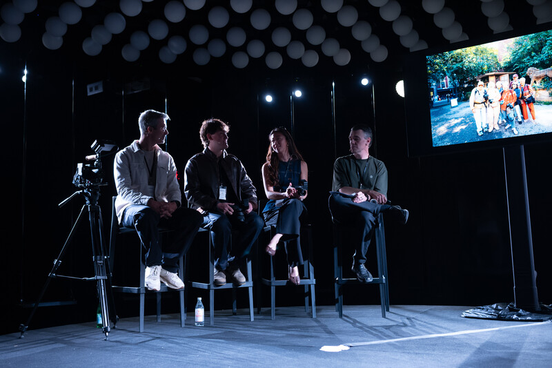 Four panelists seated on stools on a dark stage beneath glowing white orbs, with a video camera on a tripod and a projected screen on the right.
