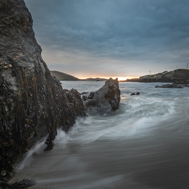 Long exposure of silky surf washing over dark rocks on a coastline at dusk, with distant hills under a cloudy sky.