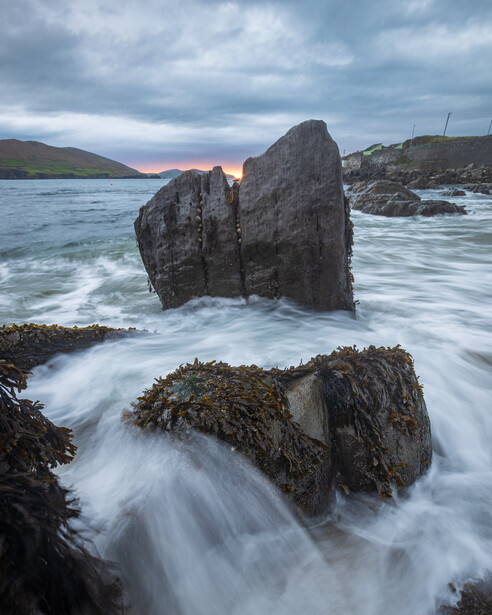 Large dark rock stack rising from churning sea with silky long-exposure water flowing around seaweed-covered boulders at dusk
