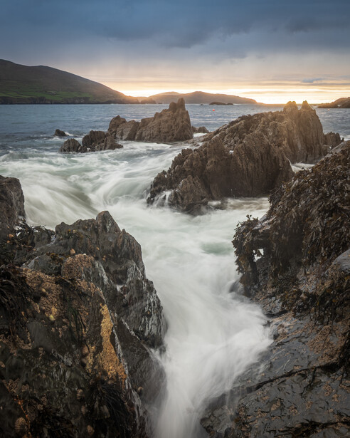 Long exposure of silky white water flowing over dark rocky coastline at sunset, with green hills and a cloudy sky in the background
