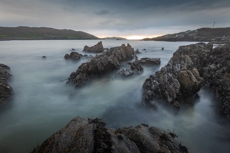 Long exposure of jagged dark rocks rising from smooth, misty sea under a moody overcast sky
