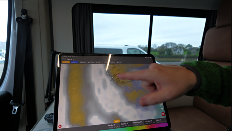 Hand pointing at a tablet showing a colorful weather forecast map inside a camper van, with an overcast window view behind it.