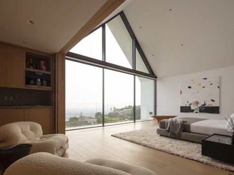 Modern bedroom with floor-to-ceiling triangular windows overlooking a coastal landscape and distant hills.