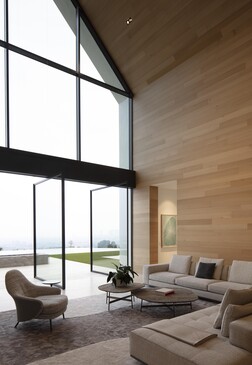 Modern living room with floor-to-ceiling windows overlooking a landscape, featuring wooden wall paneling and minimalist furnishings.