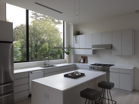 Modern kitchen interior with white cabinetry, gray countertops, island seating, and large window overlooking trees.