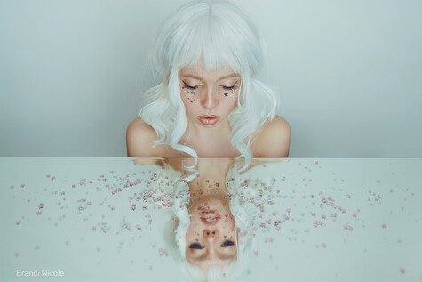 Woman with white wavy hair and glitter makeup looking down at her mirrored reflection on a white surface scattered with pink sparkles.