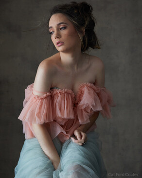 Woman in pink ruffled off-shoulder top and light blue jeans against neutral gray backdrop.