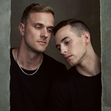 Two men in black shirts posed closely together against a neutral gray backdrop.