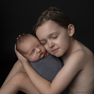 Older child embracing sleeping newborn against bare shoulder against dark background.