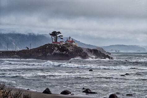 Rocky island with temple structure in turbulent ocean waters under overcast sky.