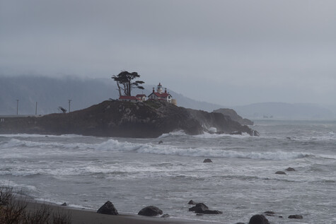 Rocky coastal island with a solitary windswept tree and building, surrounded by rough ocean waves under overcast sky.
