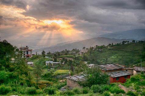 Mountain valley landscape with terraced fields, scattered buildings, and dramatic crepuscular rays breaking through clouds at sunset.