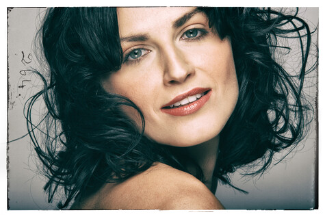 Woman with dark wavy hair and bare shoulders, shot against neutral gray background.