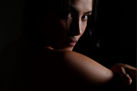 Woman with dark hair looking over shoulder against black background with dramatic side lighting.