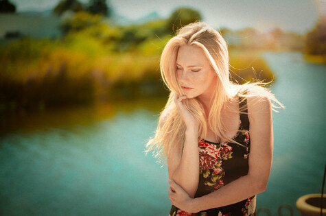 Blonde woman in floral top gazing downward beside a turquoise waterway.