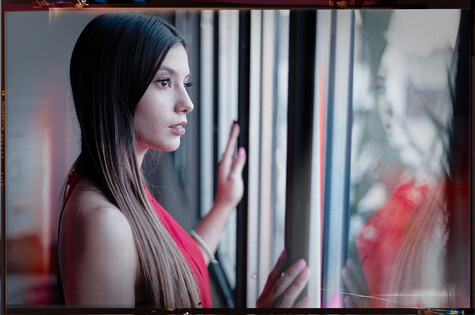 Woman with long dark hair touching a window, wearing pink, shot in profile with dramatic lighting.