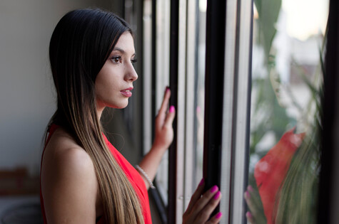 Woman with long dark hair in profile, touching a window with her hand, wearing a bright coral/pink sleeveless top.