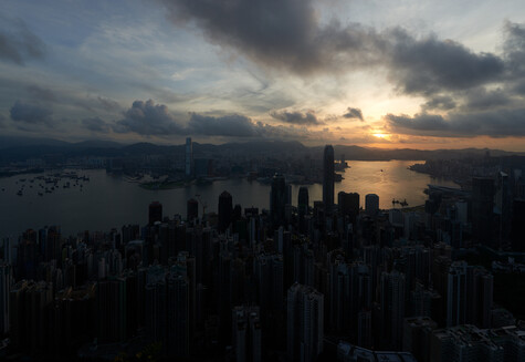 Dense urban skyline silhouetted against a golden sunset with dramatic storm clouds overhead and water visible in the foreground.