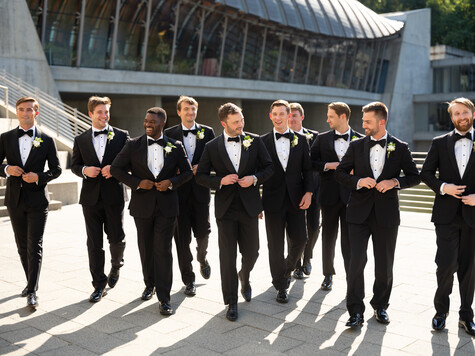 Nine men in black tuxedos walking together across a paved plaza in bright sunlight.