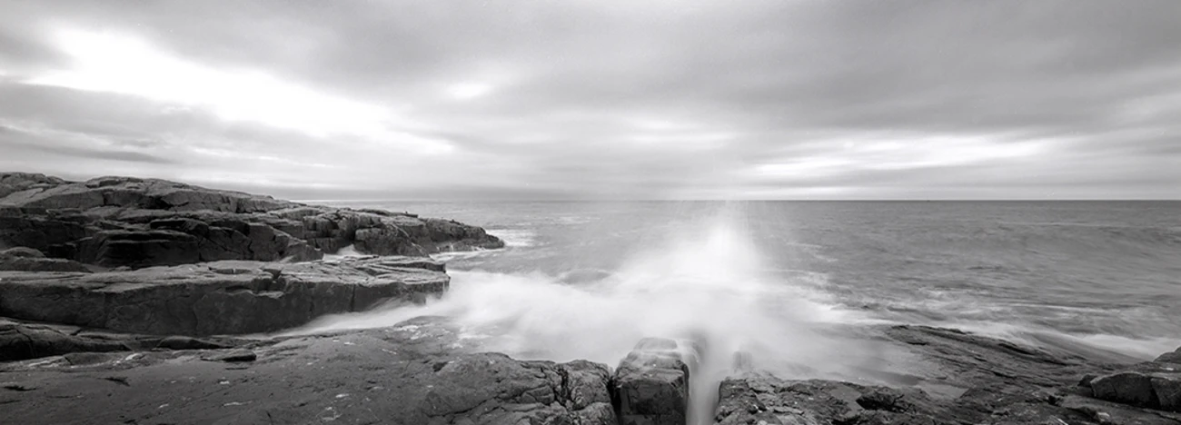 Dramatic wave crashes against dark rocky shoreline under overcast sky.