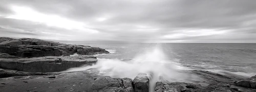 Dramatic wave crashes against dark rocky shoreline under overcast sky.