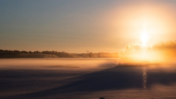 Golden sunrise over a frozen lake with forested shoreline and mist rising from the water.