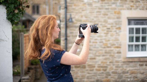 Woman with reddish-blonde hair holding a camera up to her face while standing in front of a stone building.
