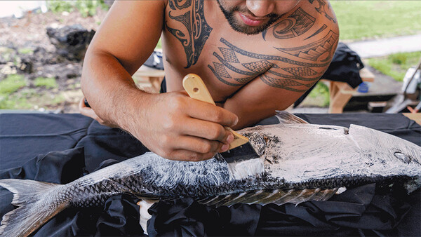 Man with traditional Polynesian tattoos examining a large fish on dark fabric outdoors.